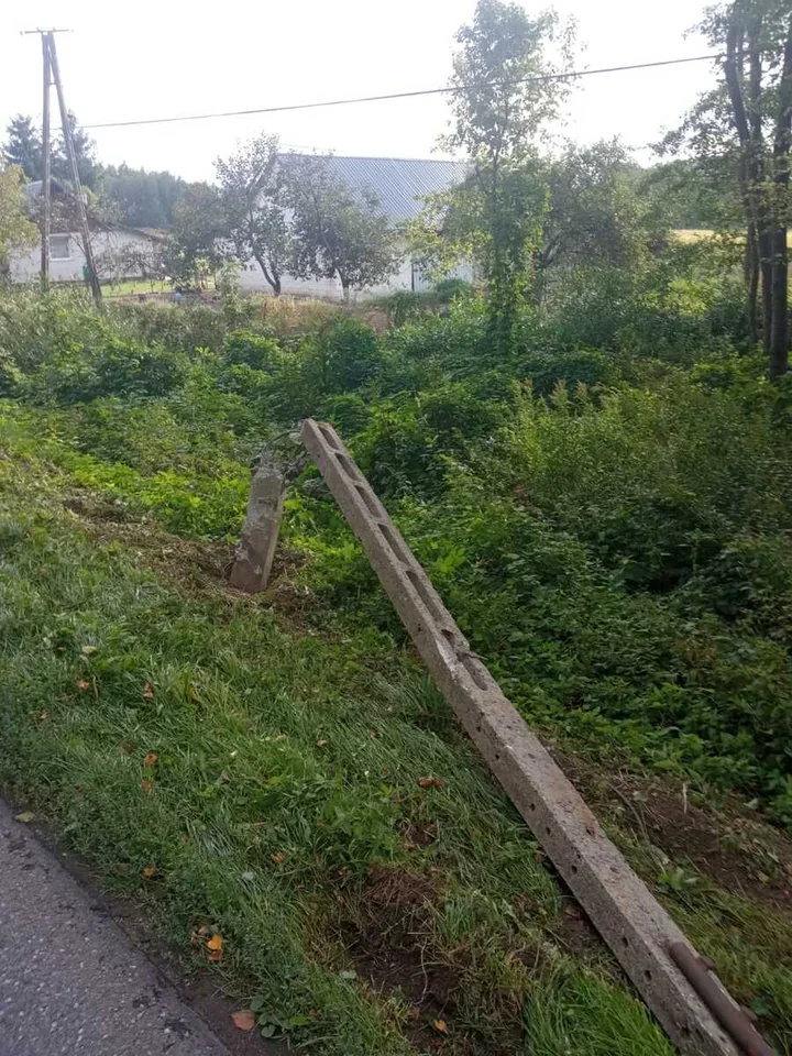 Przewrócony betonowy słup energetyczny leży na trawiastym poboczu drogi.